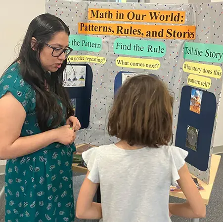 Celeste Gonzalez talking to a student in front of a poster that says math in our world, patterns, rules and stories. Find the pattern, find the rule, tell the story.