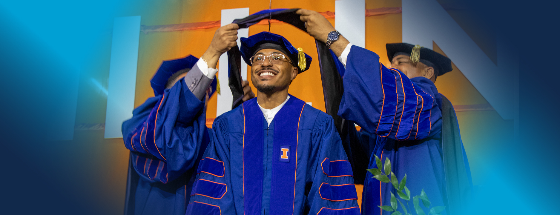 Male in a blue University of Illinois graduation gown being hooded by two male figures.