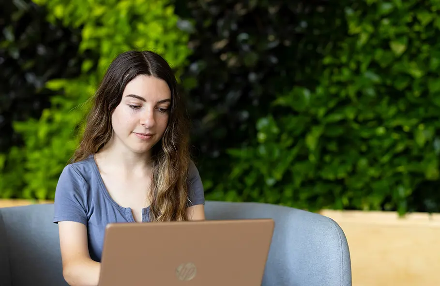 A graduate student works on a laptop in front of a plant wall