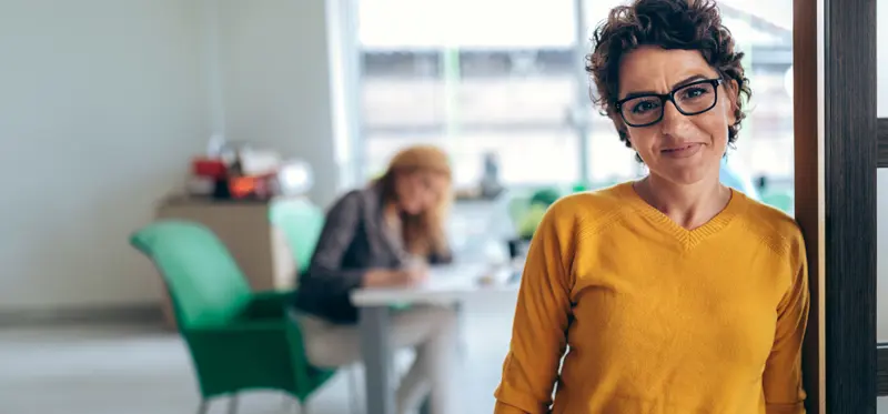 A woman in a yellow sweater with glasses smiling and standing.