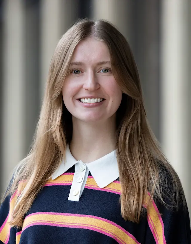 Kami smiling with a while collared and navy, yellow, and pink striped sweater.